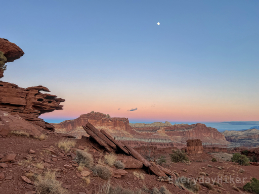 A view towards a distant cliff wall, with the jagged broken layers of a hillside of red rock framing the left. The moon may be seen well above a slightly glowing horizon.