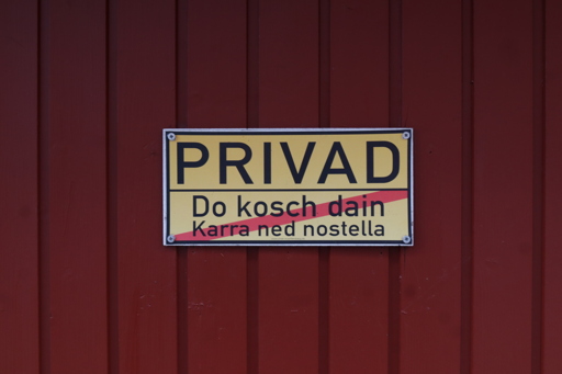 An einem roten Garagentor hängt ein gelbes Schild im Stil einer Ortsausgangstafel. In der oberen Hälfte steht "PRIVAD", in der unteren durchgestrichen "Do kosch dain Karra ned nostella".