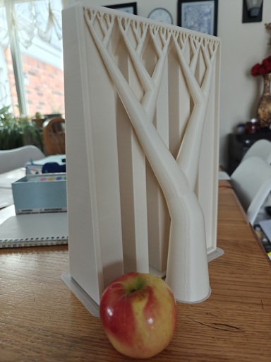 The photo shows a white 3D print sitting in front of an apple for scale. The print and apple are on a messy table. The print still has a tree support attached.
